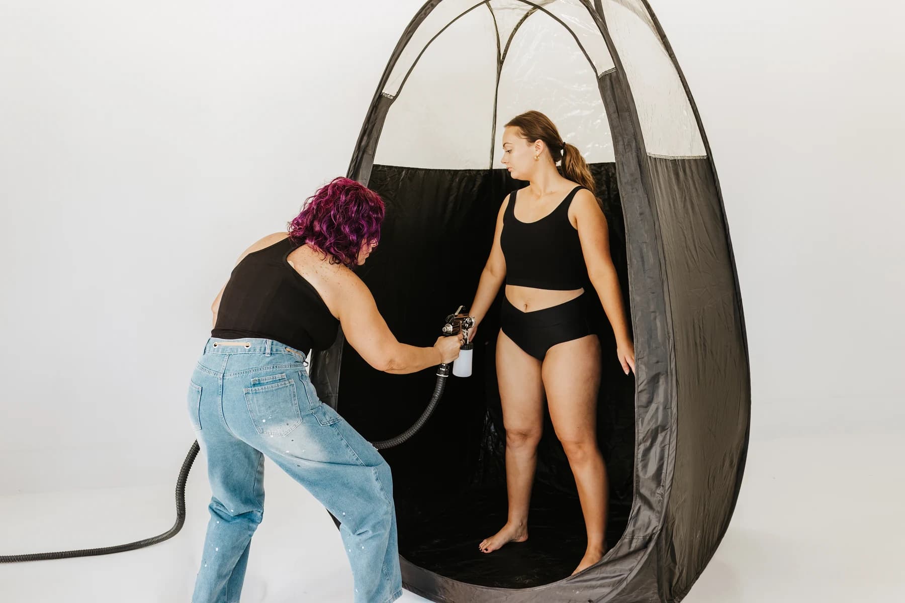 Courtney applying a spray tan to a model inside a tanning tent